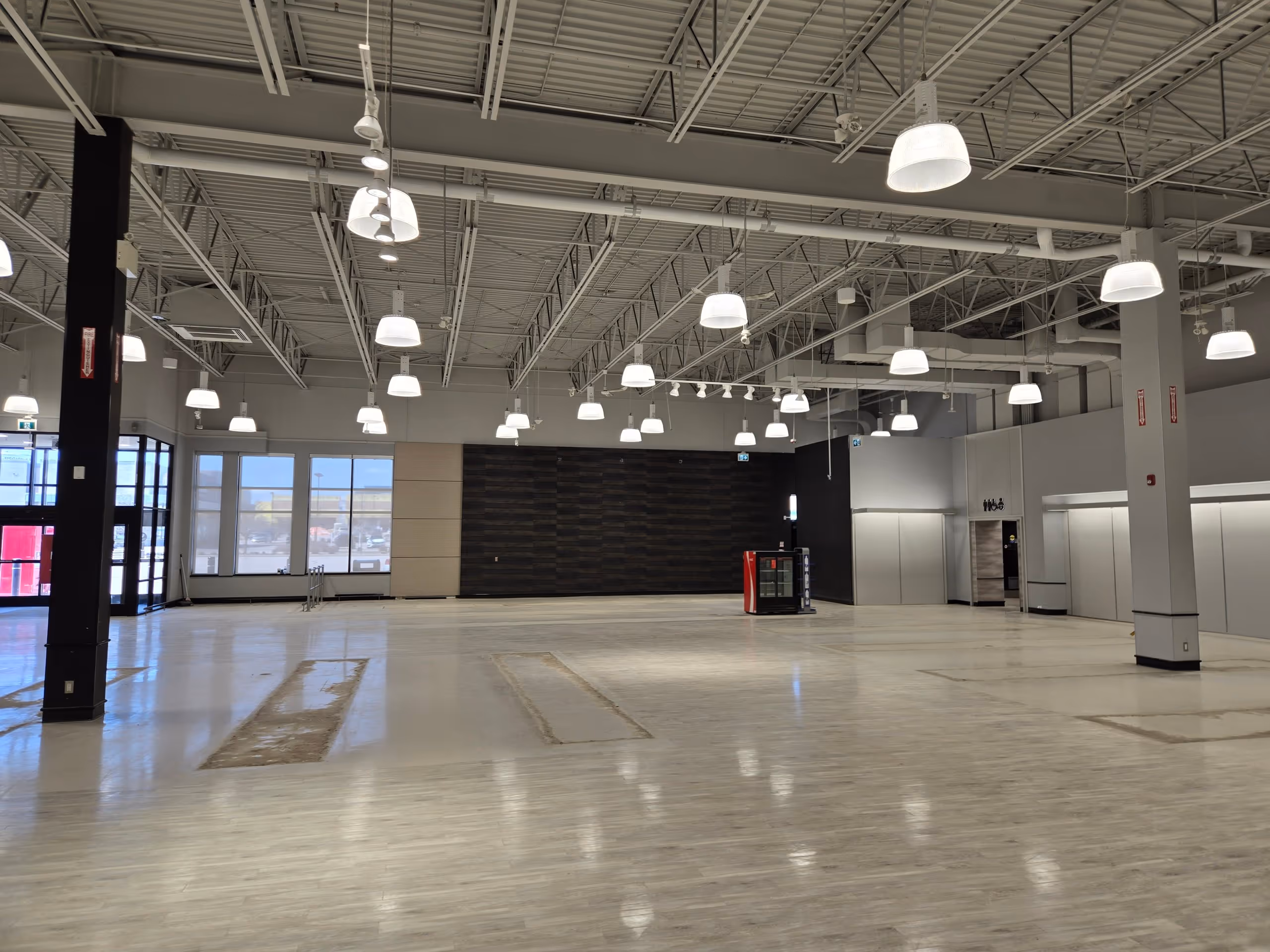 Large empty retail space with light wood flooring, high ceiling with exposed beams, many hanging lights, and a small soda vending machine near the back wall.