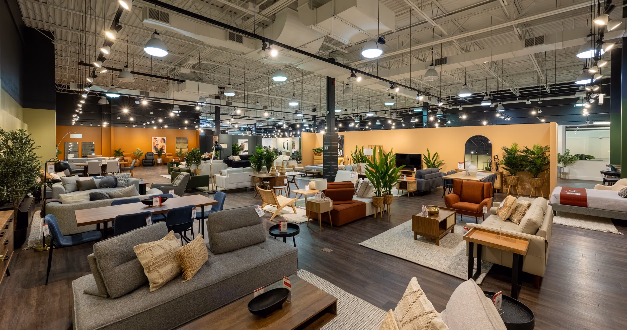 Spacious modern furniture showroom displaying sofas, armchairs, dining tables, beds, and decorative plants under bright ceiling lights.