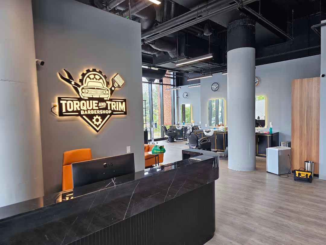 Modern barbershop interior with a black marble reception desk, orange chair, illuminated Torque and Trim Barbershop sign on the wall, and styling stations with chairs and mirrors.