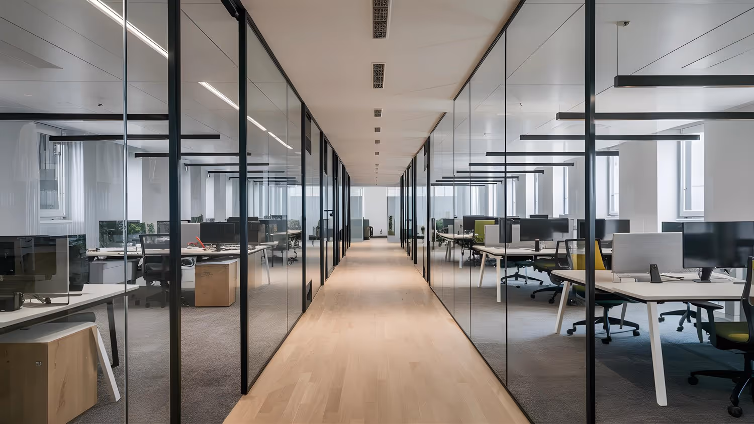 Modern office corridor with glass-walled rooms featuring desks, computer monitors, and office chairs.