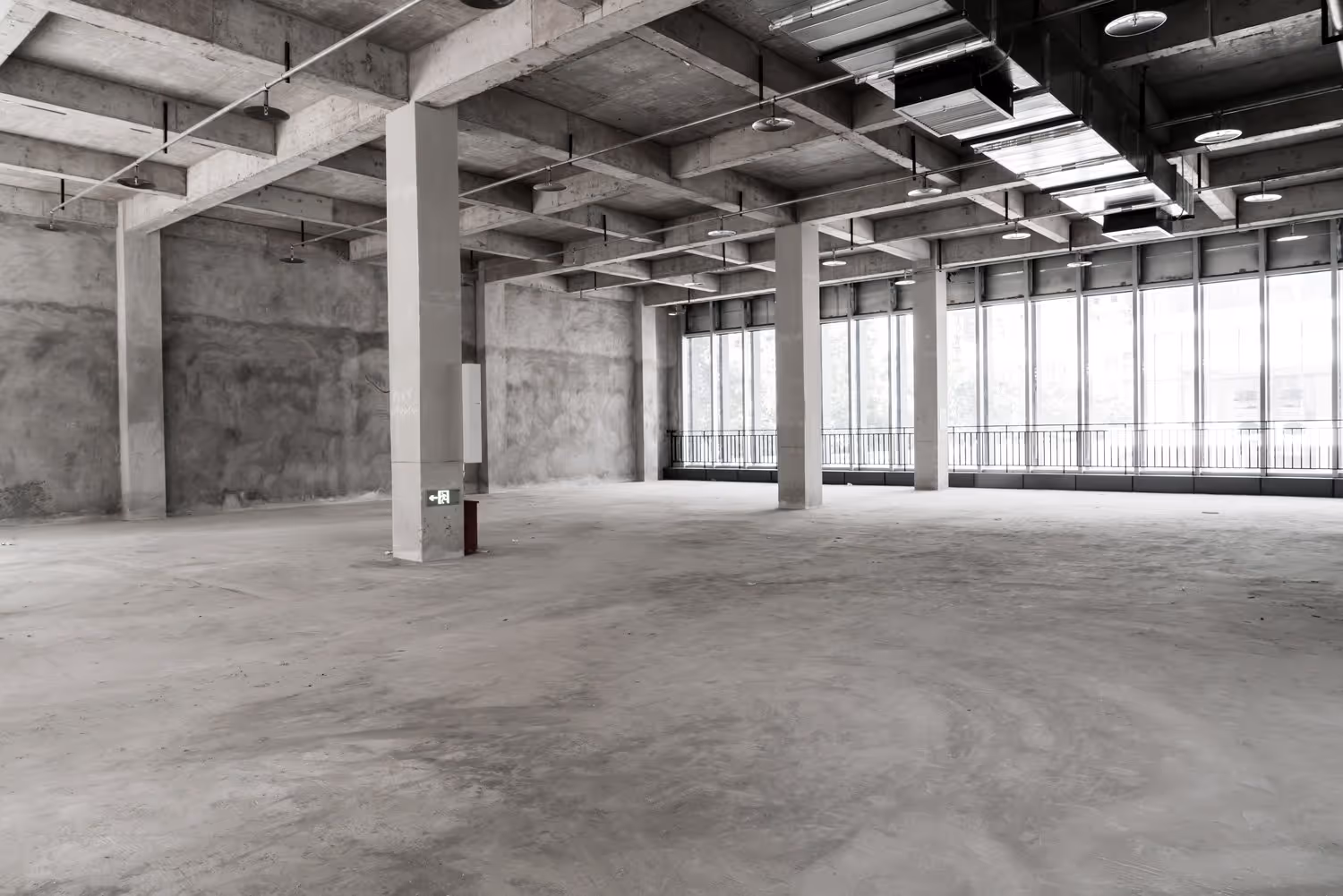 Large empty industrial space with concrete floors, ceiling beams, and tall windows along one wall.