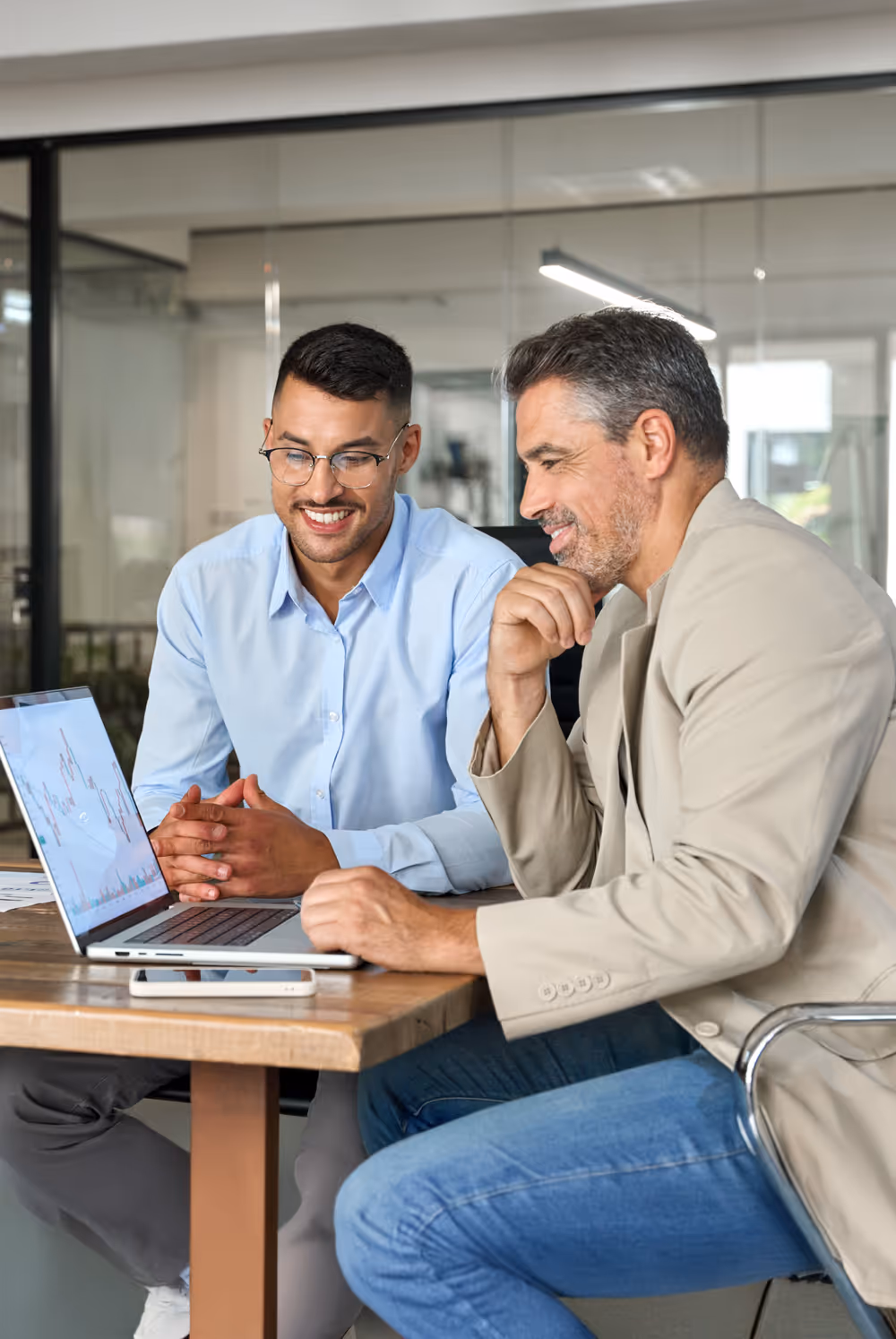 Two businessmen sitting at a table looking at a laptop screen displaying financial charts and discussing.