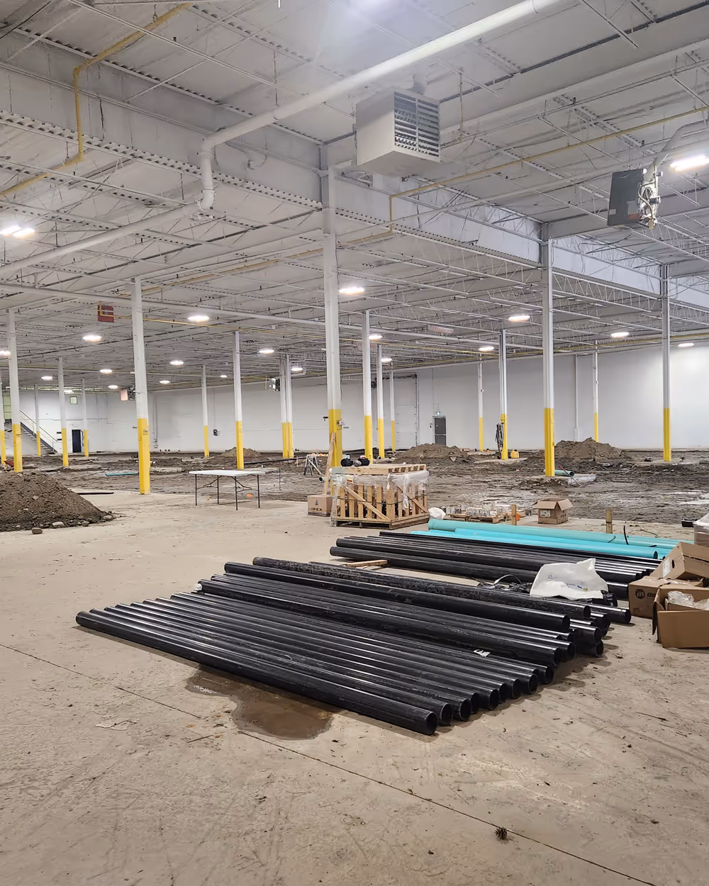 Interior of a large industrial warehouse under construction with stacks of black and turquoise pipes on the floor.
