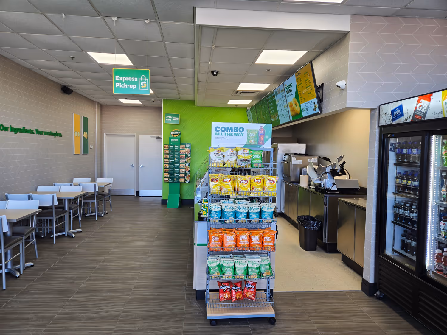 Interior of a Subway restaurant with snack display of chips, a refrigerated drink cooler, dining tables, and digital menu boards over the counter.