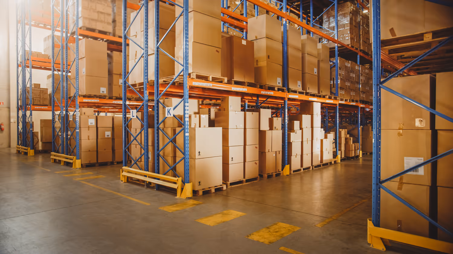 Industrial warehouse interior with tall blue and orange metal racks filled with stacked cardboard boxes on wooden pallets.