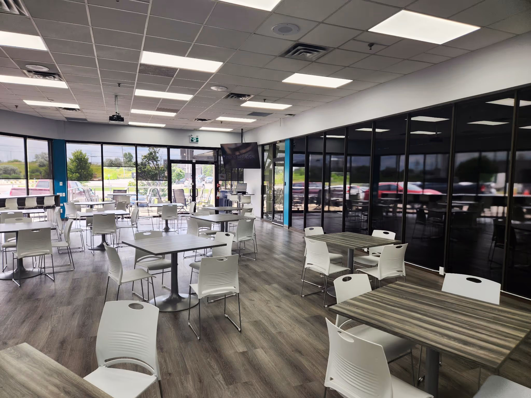 Modern office break room with multiple tables and white chairs, large windows, and wood-patterned flooring.