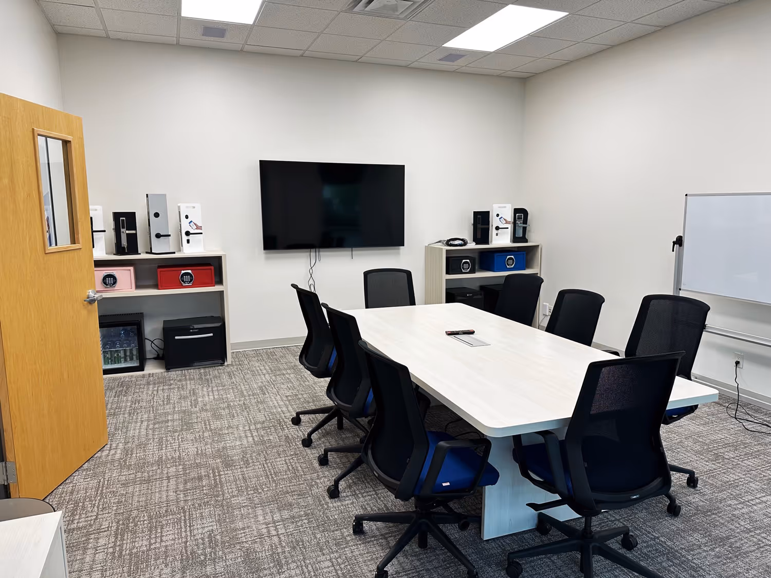 Modern conference room with a rectangular white table surrounded by eight black mesh office chairs, a wall-mounted flat screen TV, two shelves with locks and devices, a whiteboard, and a wooden door with a window.