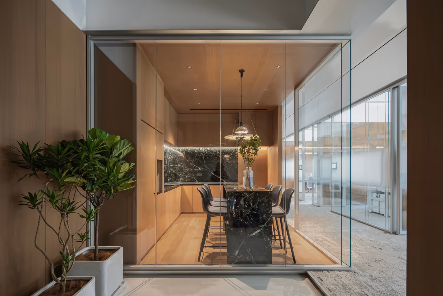 Modern office kitchen with wooden cabinets, black marble island, six bar stools, and indoor plants in white pots.
