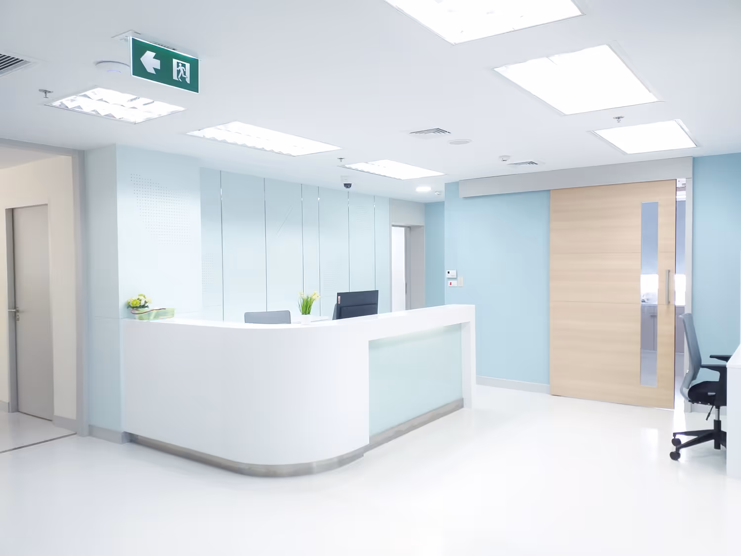 Bright modern reception area with white desk, office chairs, light blue walls, and overhead fluorescent lights.
