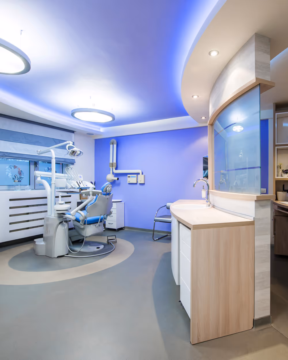 Modern dental clinic room with dental chair, equipment, blue accent wall, and a curved countertop with sink.
