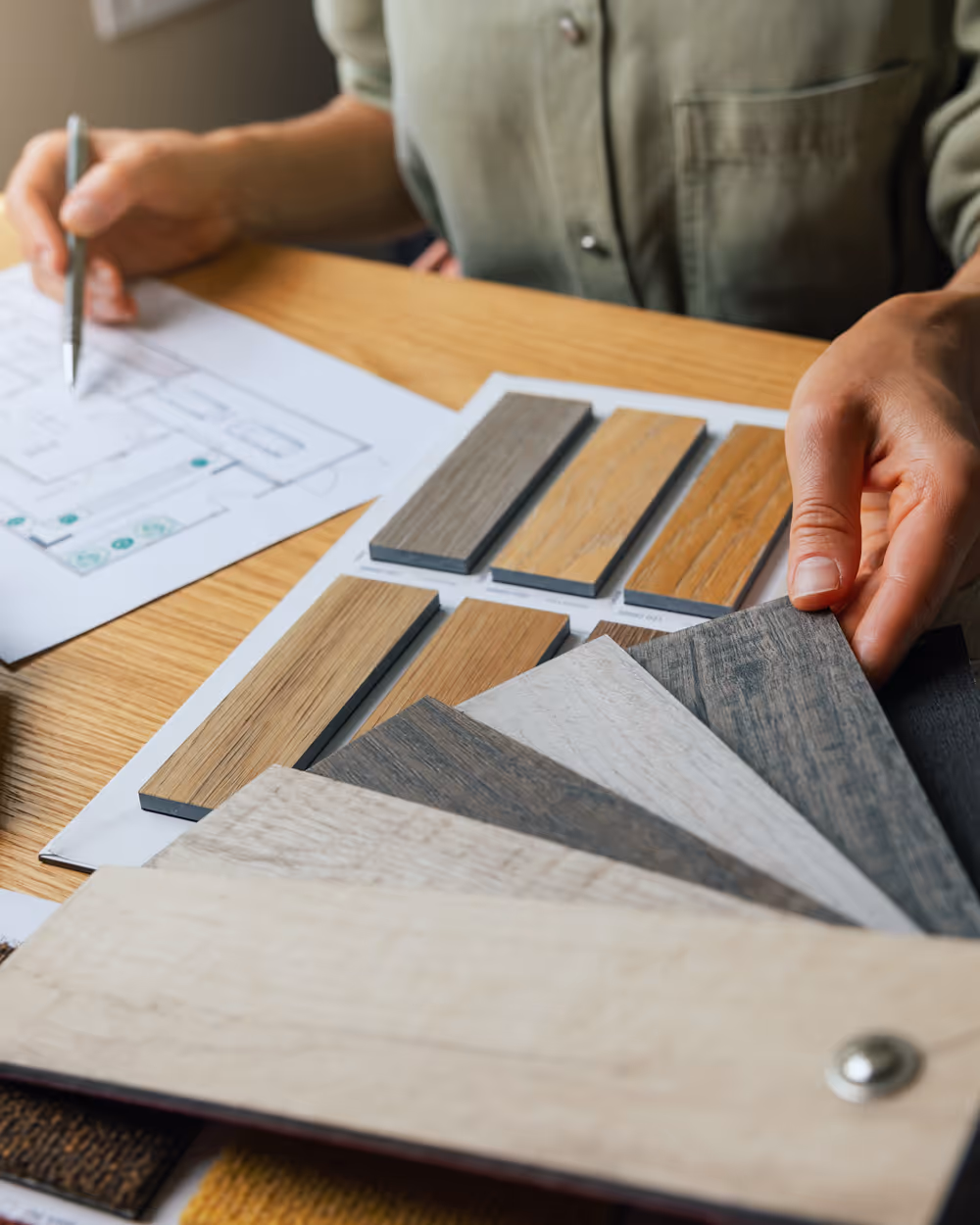 Person comparing various wood flooring samples and reviewing a floor plan on a wooden table.
