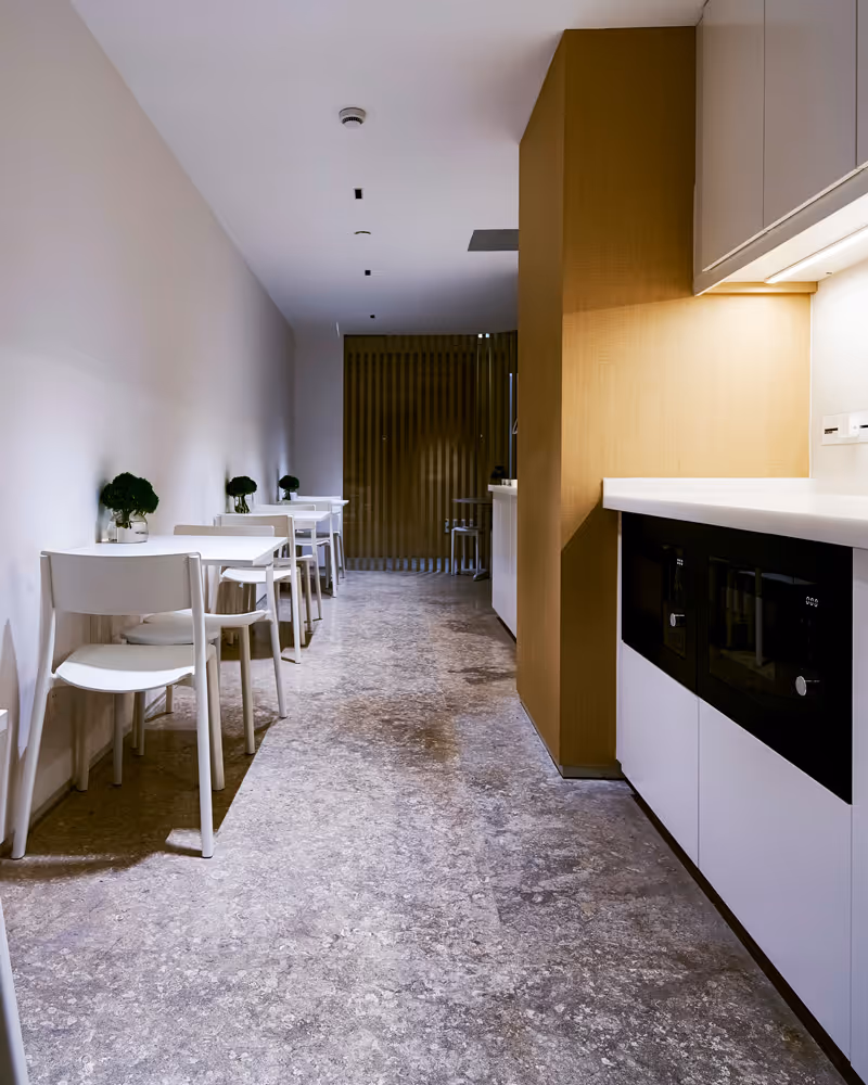 Minimalist dining area with white tables and chairs lined along a white wall, small green plants on each table, and a modern kitchen area with white countertops and wooden paneling.