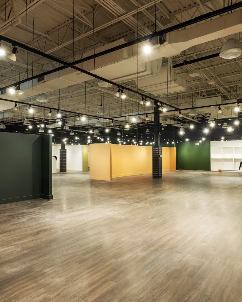 Spacious empty room with wood flooring, green and yellow partition walls, and numerous ceiling lights.