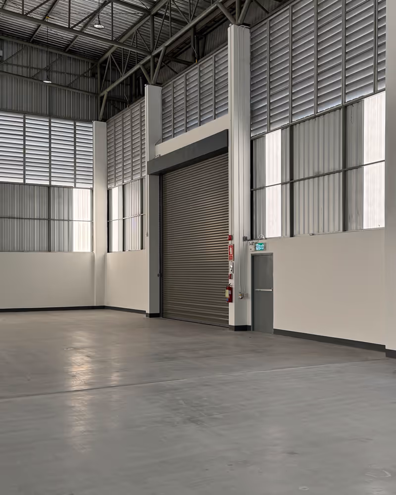 Spacious empty warehouse interior with concrete floor, large metal roll-up door, and tall white walls with horizontal windows near the ceiling.