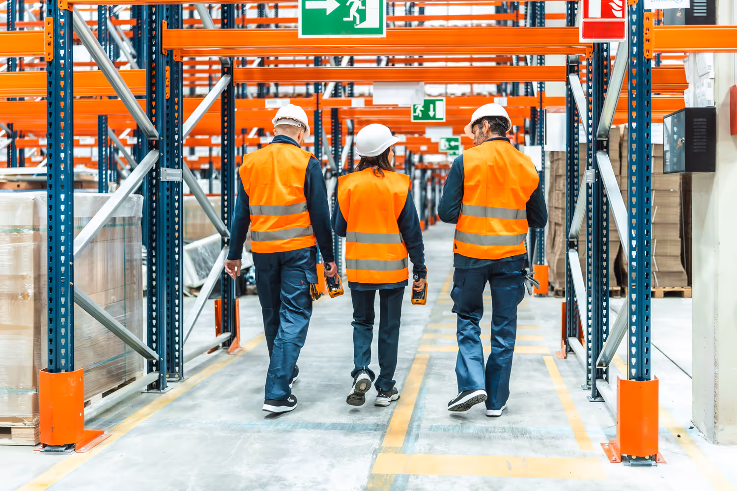 Three warehouse workers wearing orange safety vests and white helmets walking down an aisle between storage racks.