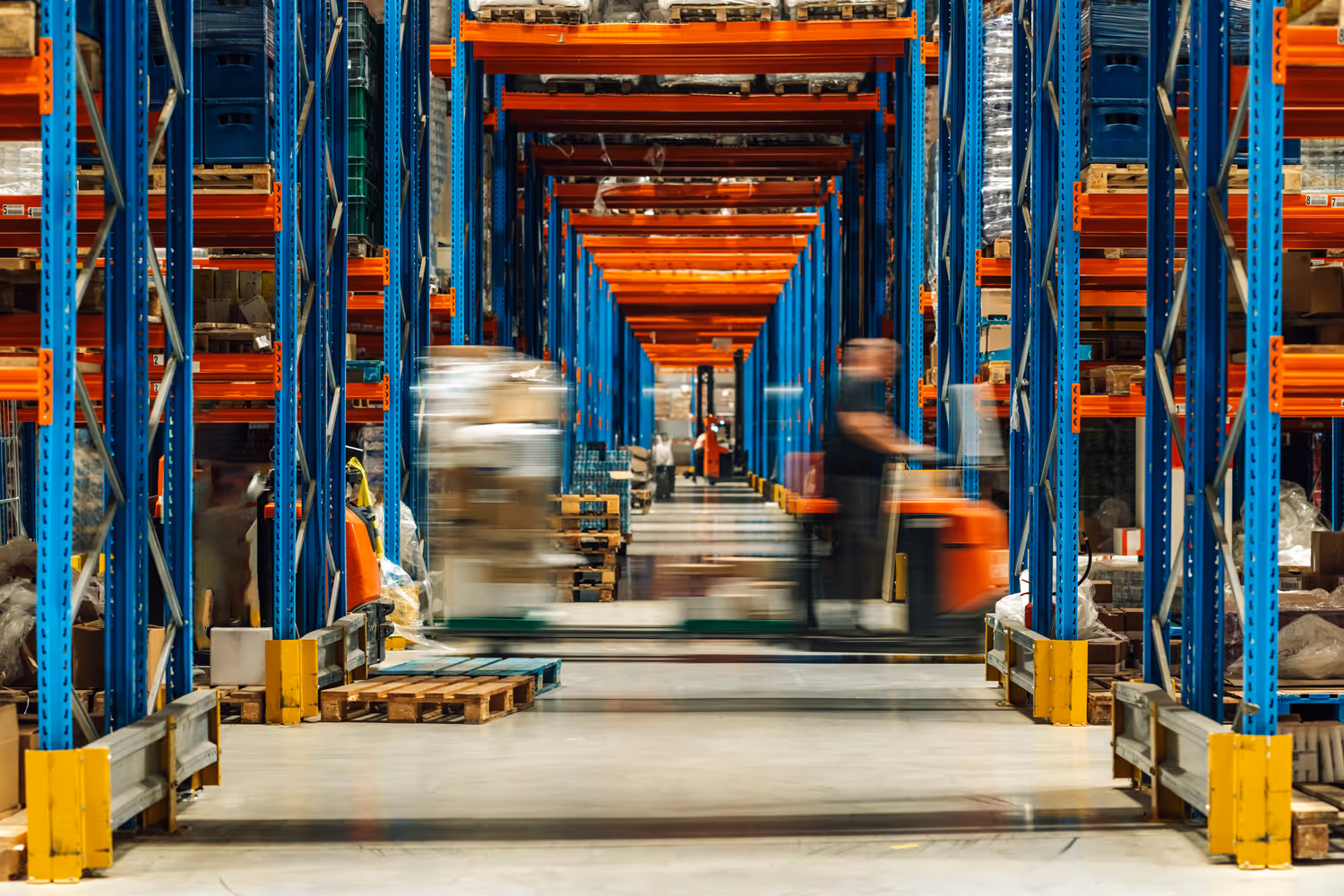 Blurred workers moving pallets in a large warehouse with blue and orange storage racks.