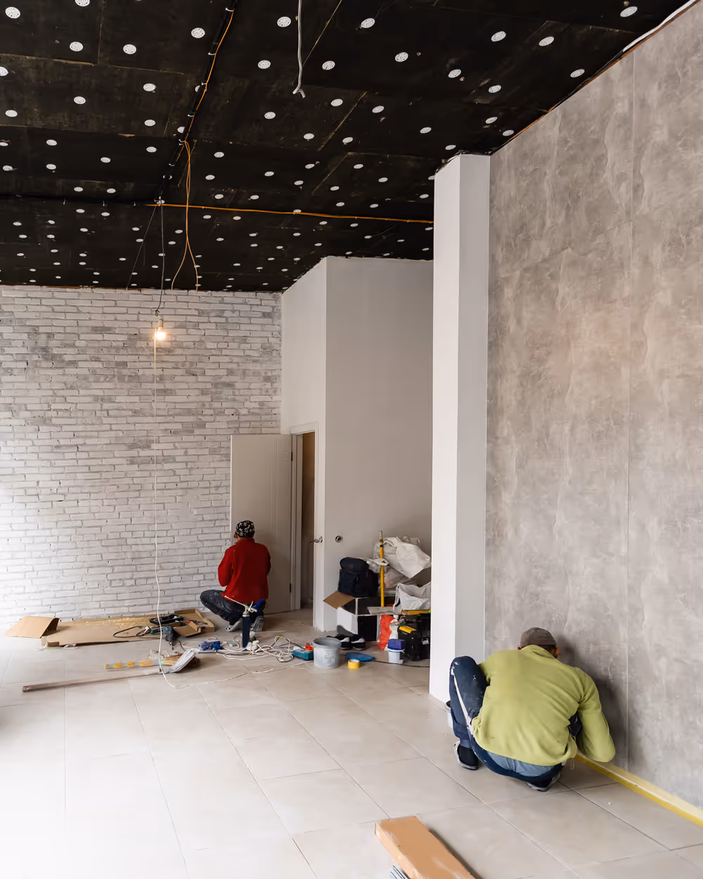 Two workers renovating a room with one measuring the wall and the other working near a white brick wall, surrounded by tools and construction materials.