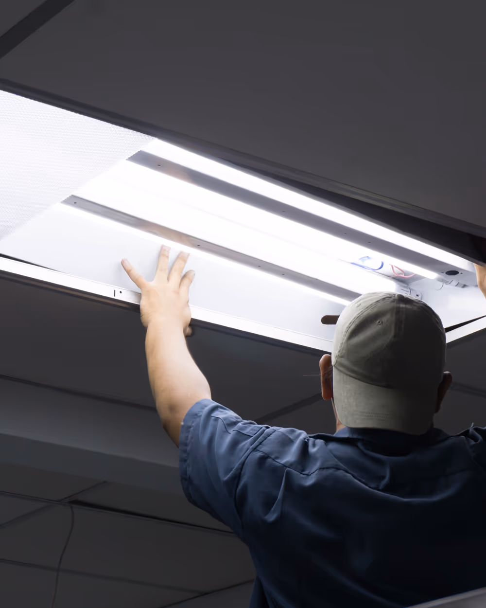 Man wearing a cap replacing a ceiling light panel with bright fluorescent tubes.