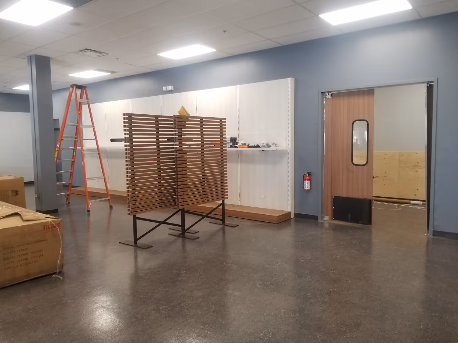 Empty commercial space with a wooden display rack, an orange ladder, cardboard boxes, and a wooden door leading to another room.