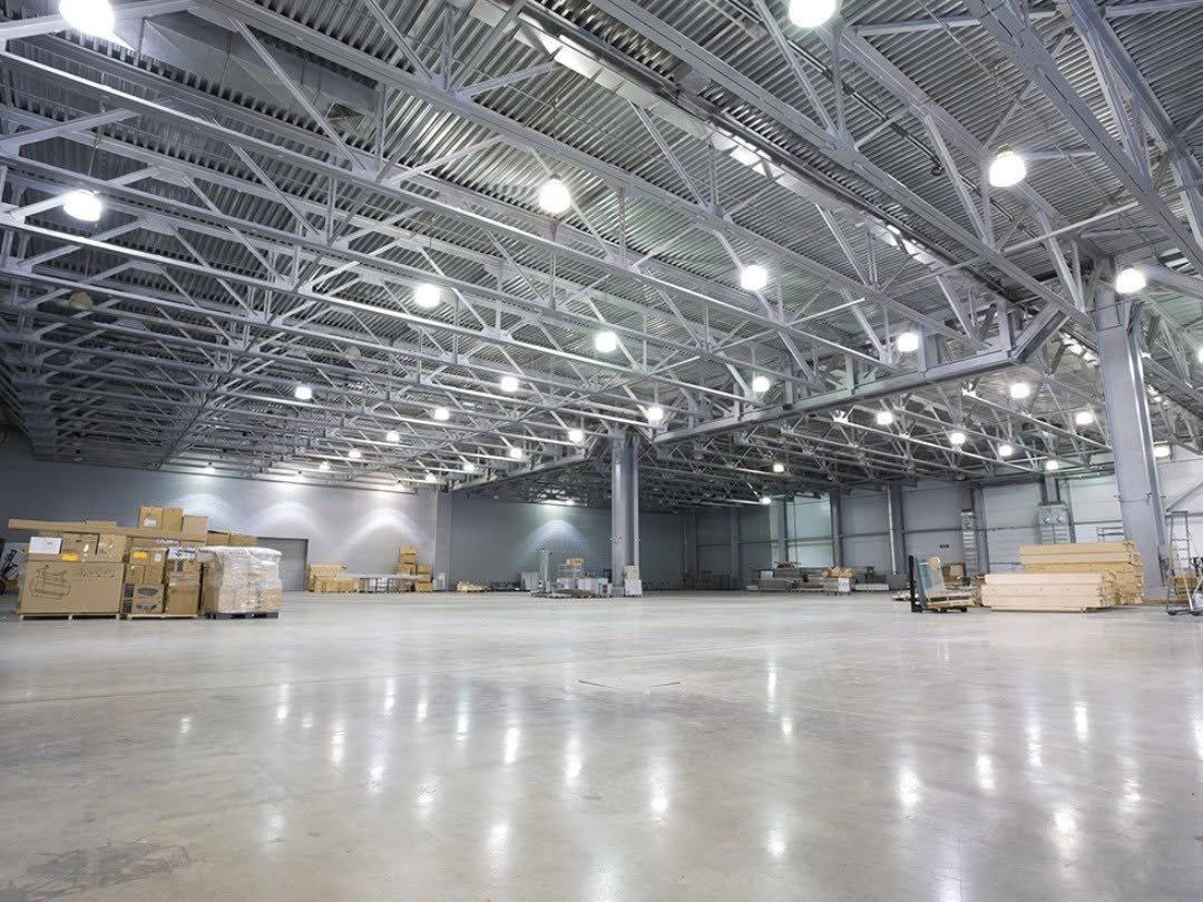 Large, bright empty warehouse with high ceiling and stacked cardboard boxes and pallets along the walls.