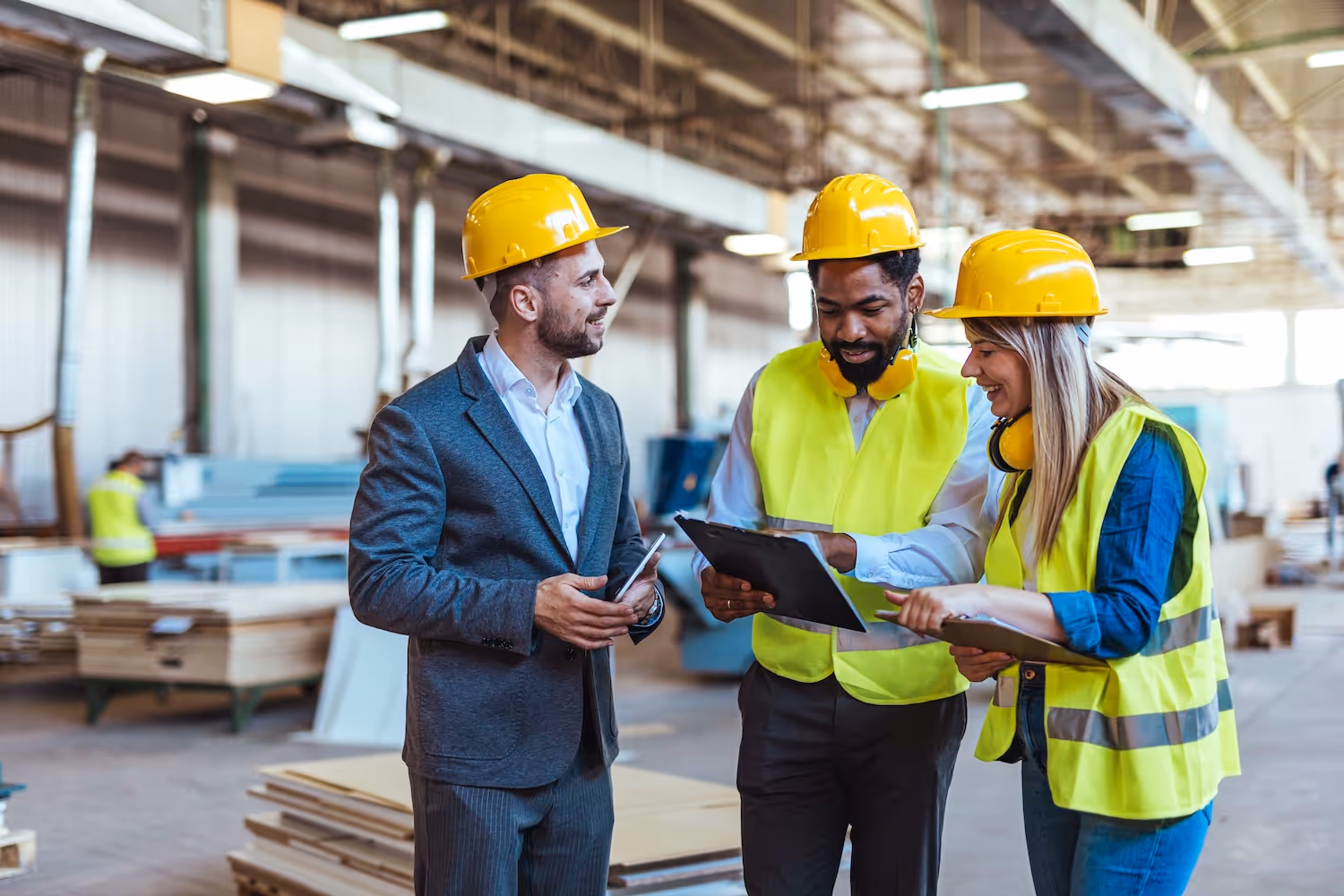 Three construction workers wearing yellow hard hats and reflective vests discussing papers in a large warehouse.