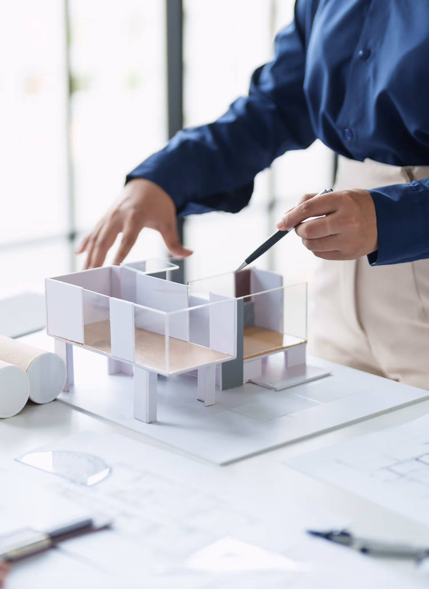 Hands of a person in a blue shirt pointing with a pen at a miniature architectural model on a table with plans and drawings.