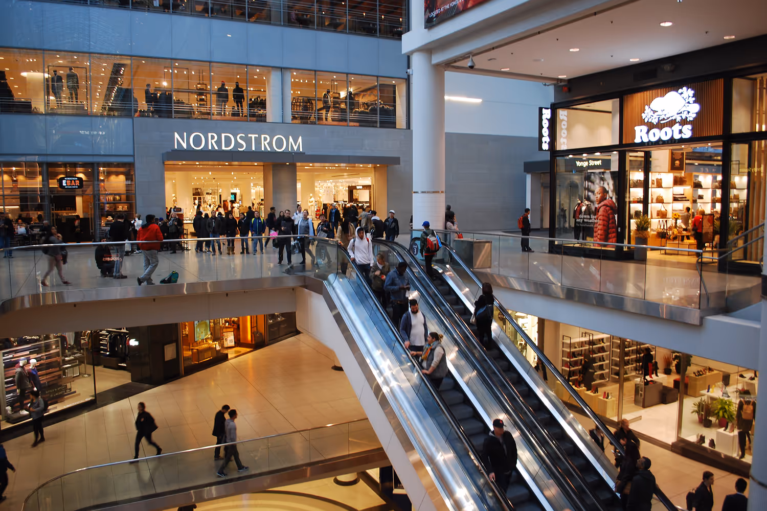 Interior view of a shopping mall with people on escalators and shoppers walking near Nordstrom and Roots stores.