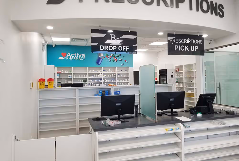 Interior of a modern pharmacy with white counters, shelves stocked with medication, and signs for prescription drop off and pick up.