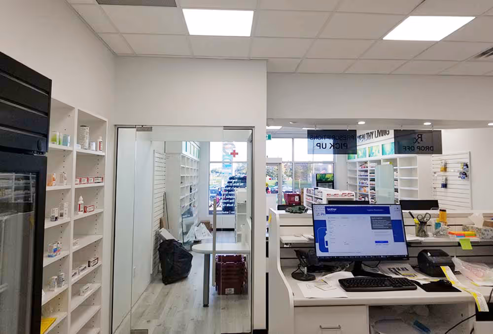 Interior of a pharmacy with white shelves containing medicine, a glass door, and a counter with a computer and office supplies.