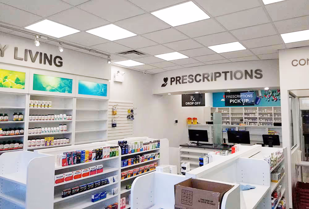 Bright and organized pharmacy interior showing shelves stocked with medication, counters with computer monitors, and signs for prescriptions drop off and pick up.