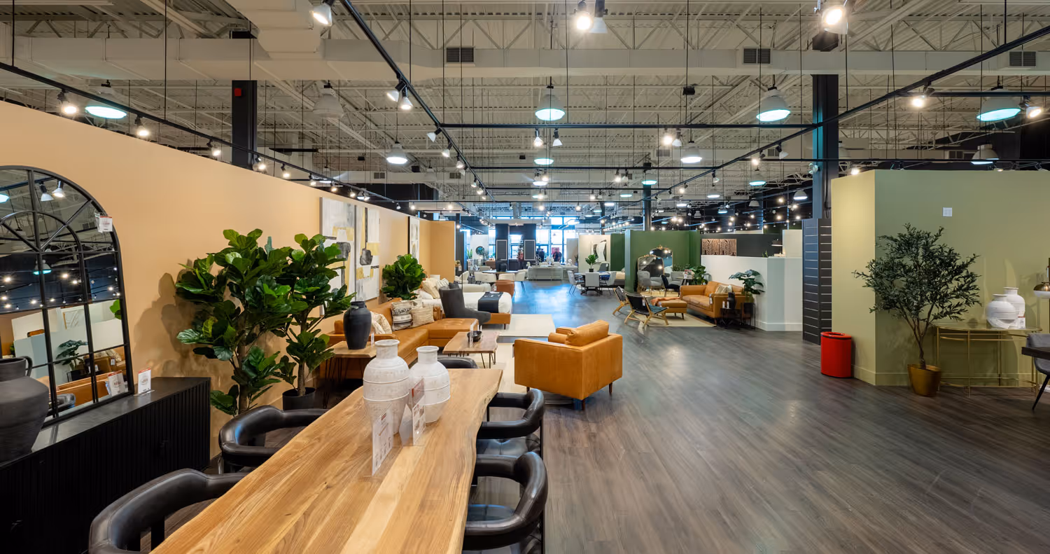 Spacious furniture showroom with wooden dining table, leather sofas, decorative plants, and modern lighting.