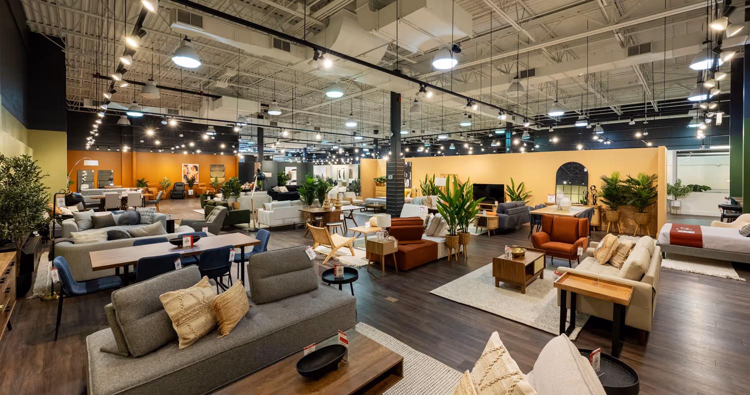 Spacious modern furniture showroom featuring multiple living room sets, dining tables, decorative plants, and bright overhead lighting.