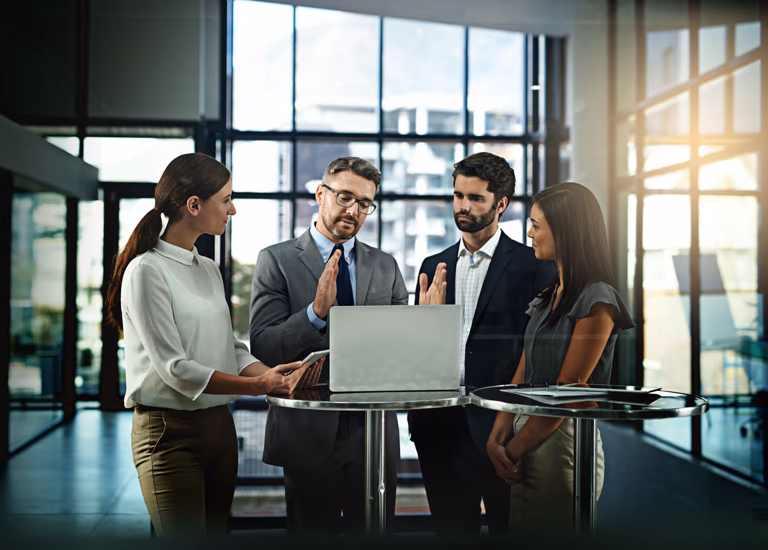 Four business professionals standing around a high table with a laptop, engaged in a discussion in a modern office.