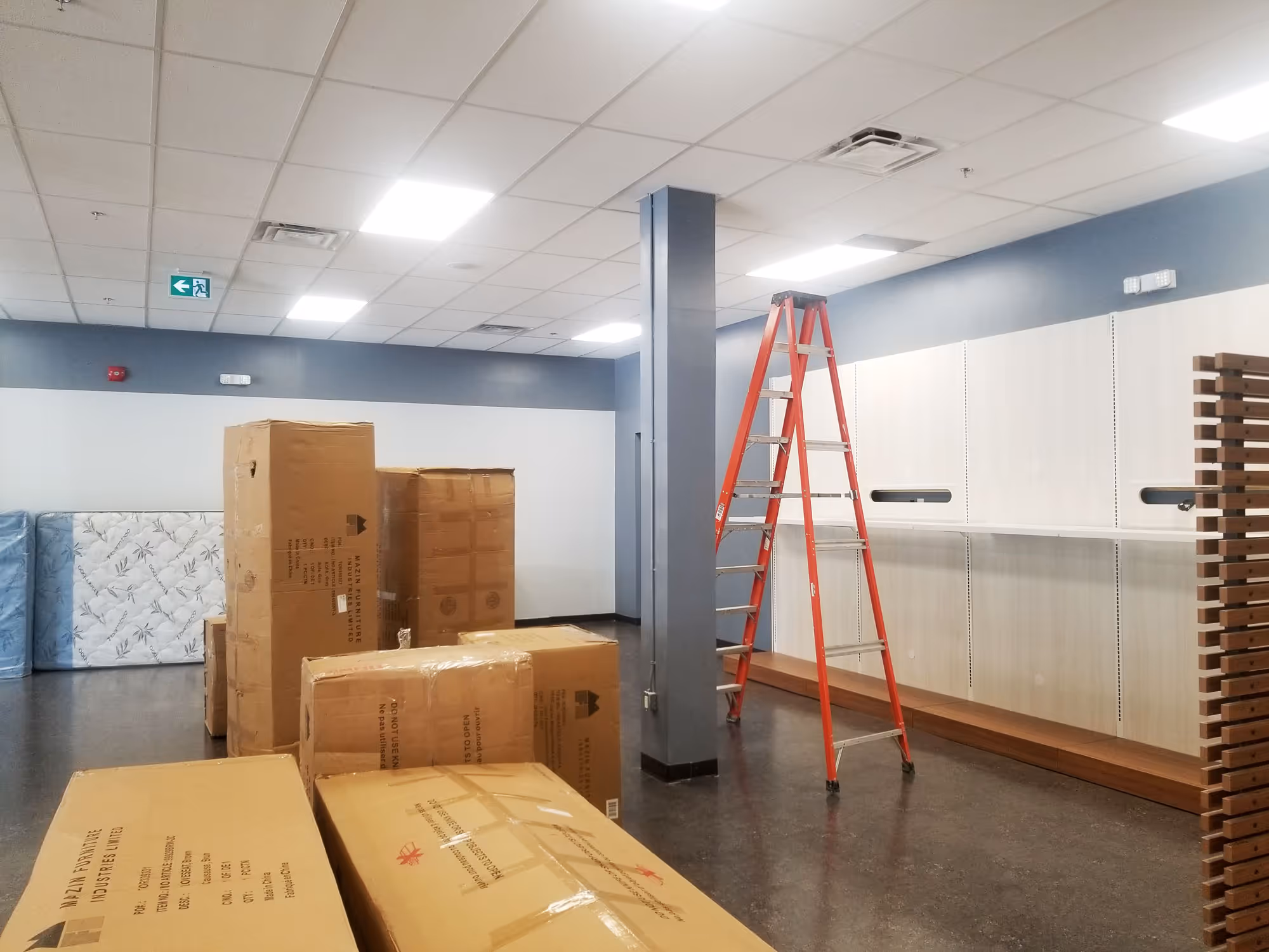 Empty room with stacked cardboard boxes, mattresses, and a red ladder near white wall shelves.