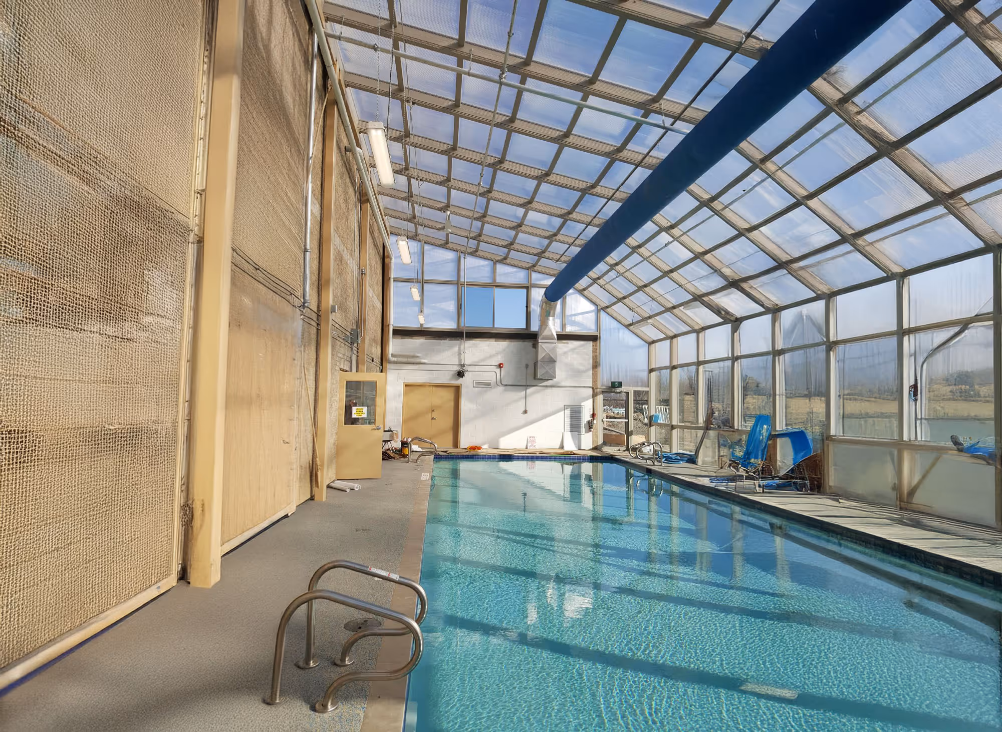 Indoor swimming pool with clear water under a glass roof and beige walls with netting on the left side.