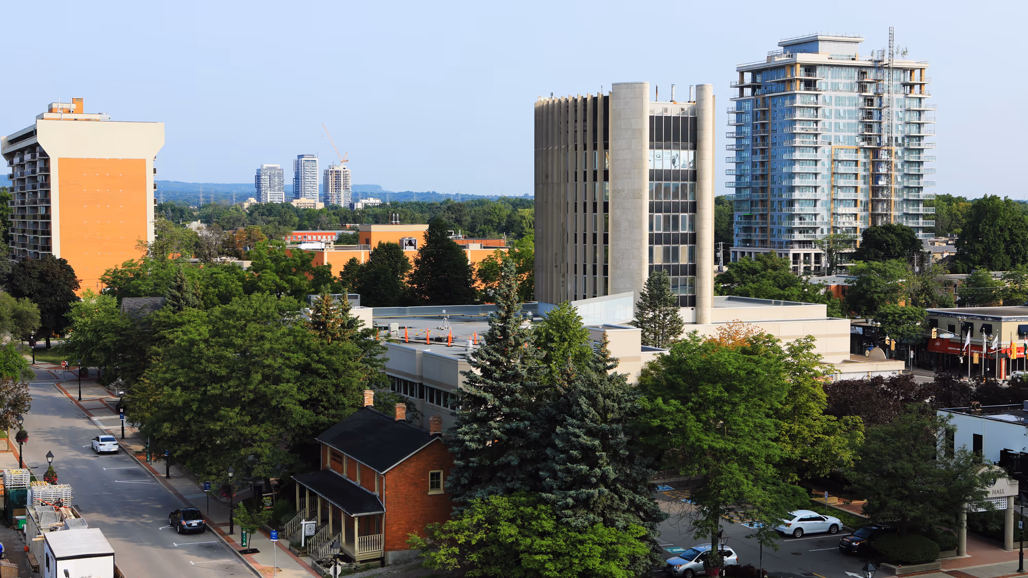 Cityscape showing a mix of modern high-rise buildings and older low-rise structures surrounded by green trees under a clear sky.