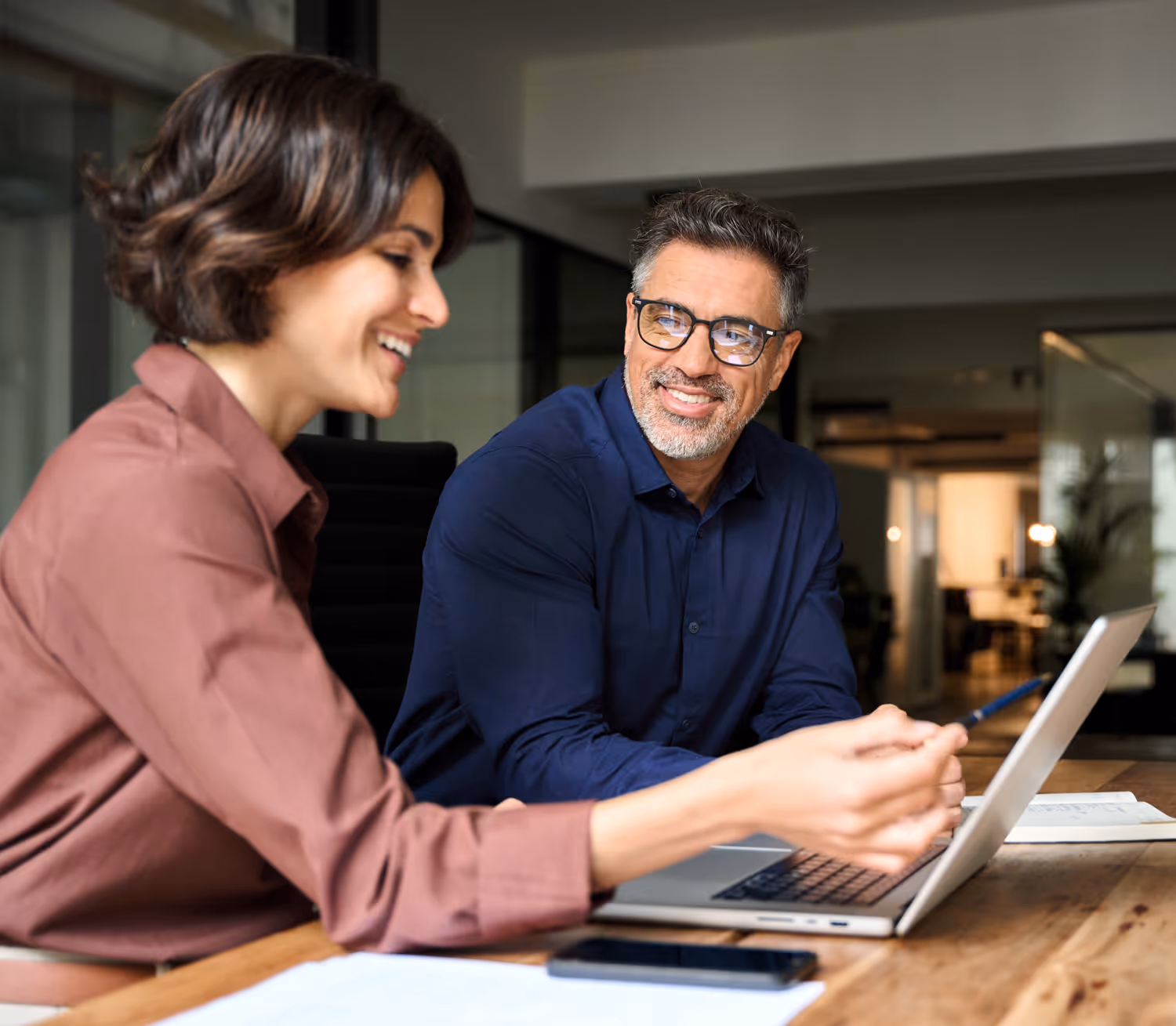 Two colleagues smiling and discussing work while looking at a laptop in a modern office.
