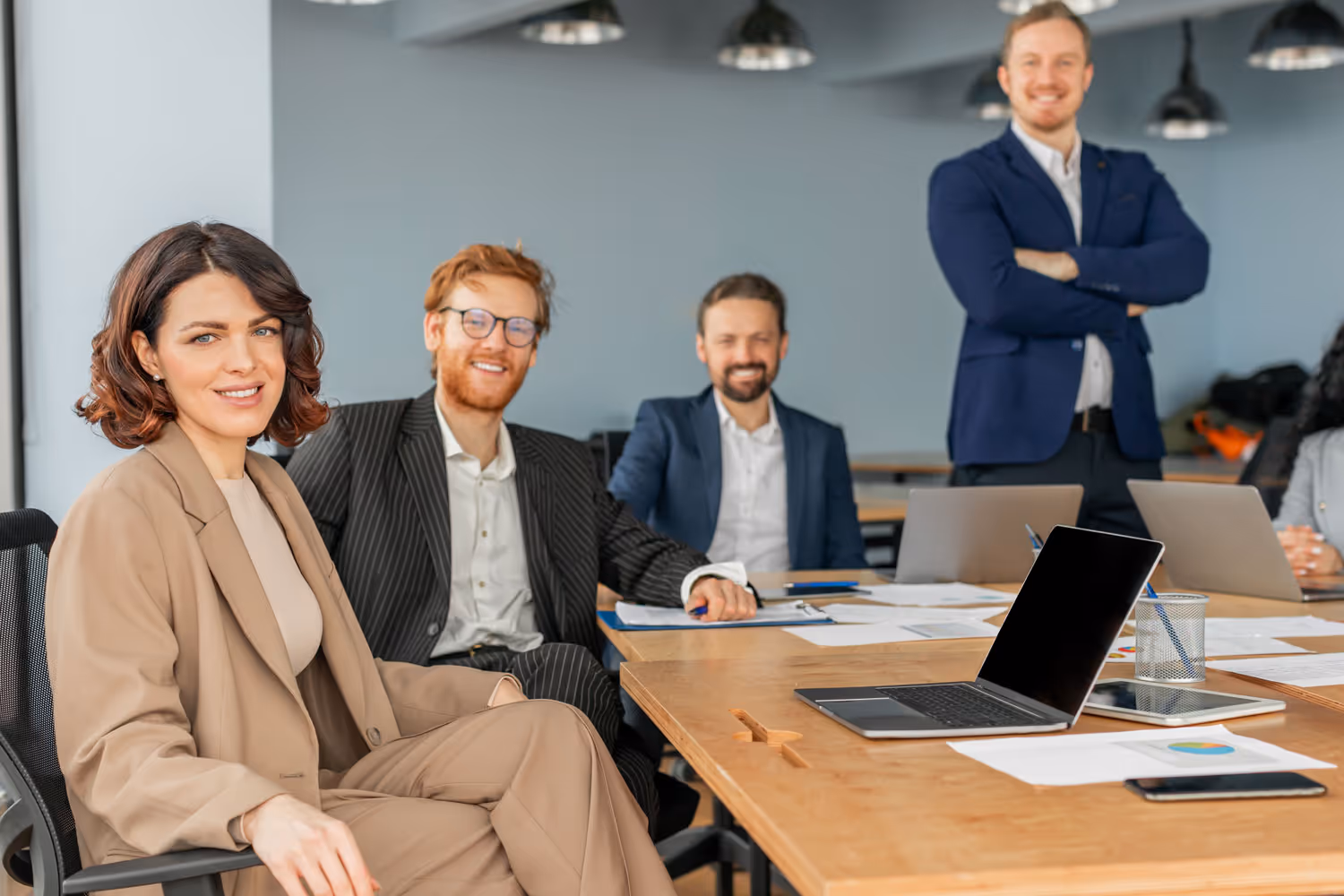 Four business professionals in a modern office, three seated at a table with laptops and documents, one standing with arms crossed, all smiling.