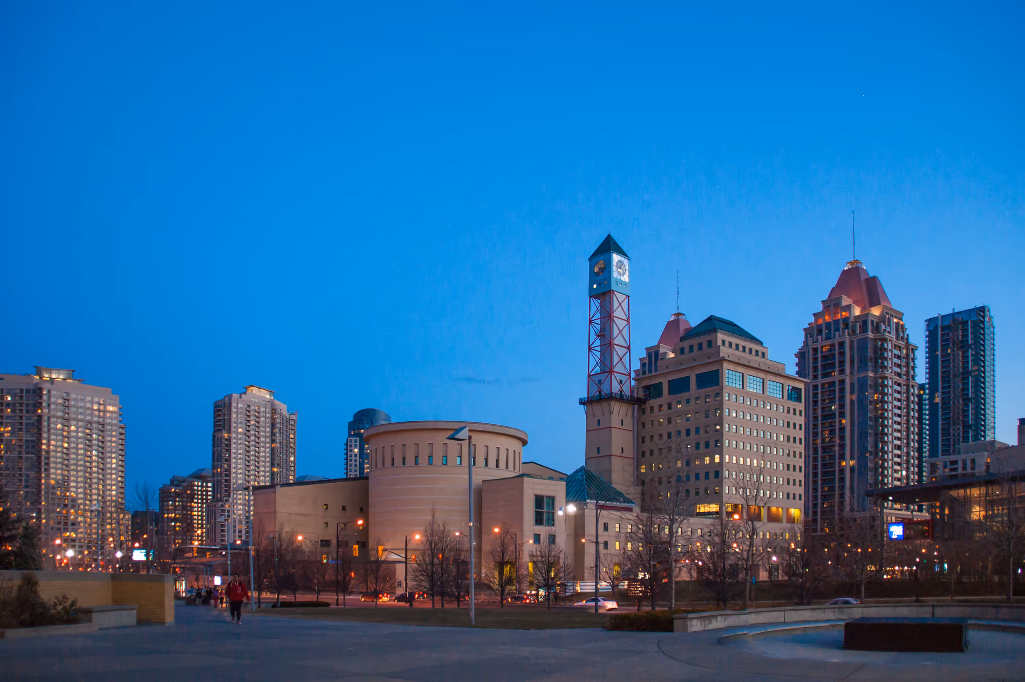 Evening view of Mississauga city skyline with illuminated buildings and a prominent clock tower against a clear blue sky.