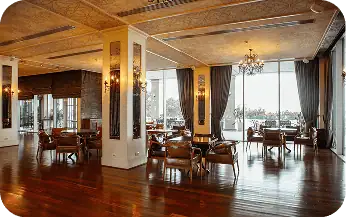 Spacious restaurant interior with wooden floors, large windows with dark curtains, and elegant chandeliers.