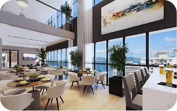 Bright modern hotel dining area with large windows overlooking a cityscape and mountains, featuring round tables with chairs and a bar counter.