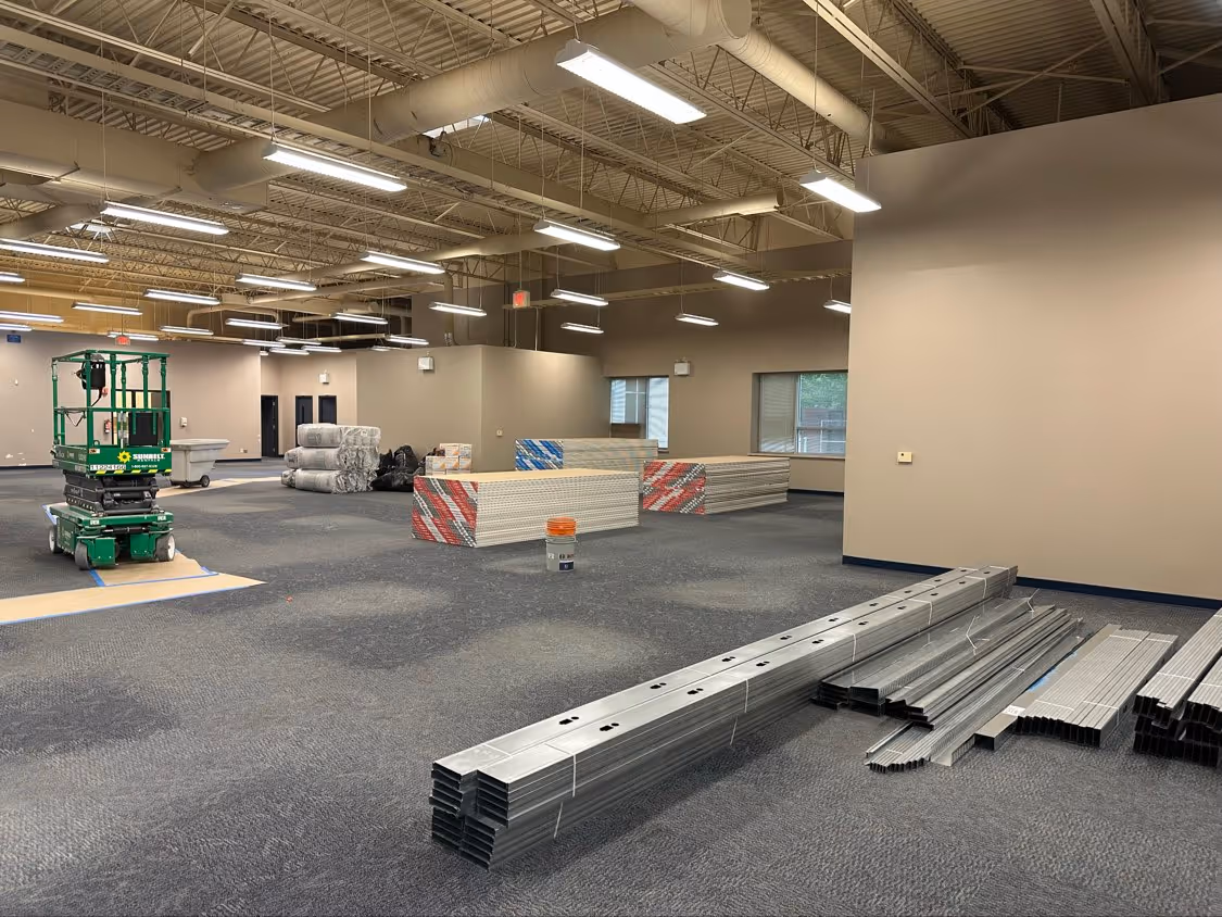 Interior of a large room under renovation with construction materials and a green scissor lift on a carpeted floor.