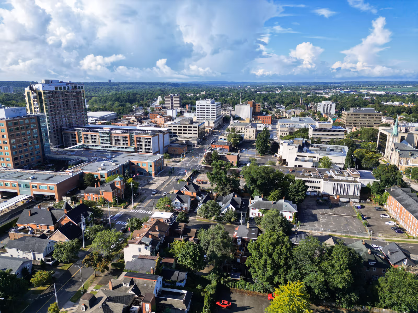 Aerial view of a cityscape with a mix of residential houses, trees, and mid-rise office buildings under a partly cloudy blue sky.