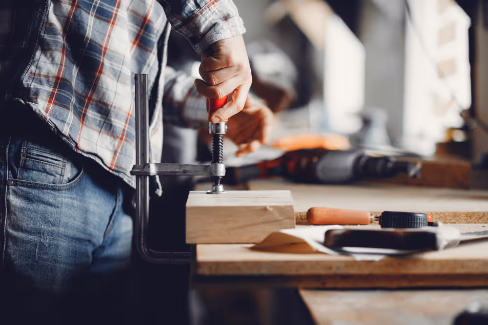 Eine Person spannt ein Holzstück mit einer Schraubzwinge in einer Werkstatt fest.