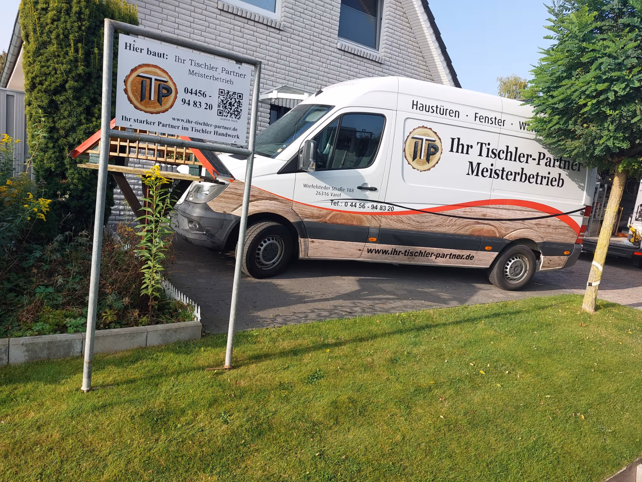 Weißer Transporter mit Werbung für 'Ihr Tischler-Partner Meisterbetrieb' neben einem Haus und einem Schild mit Kontaktdaten.