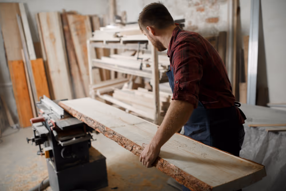 Ein Tischler schiebt ein großes unbehandeltes Holzbrett in einer Werkstatt mit Holzregalen im Hintergrund.