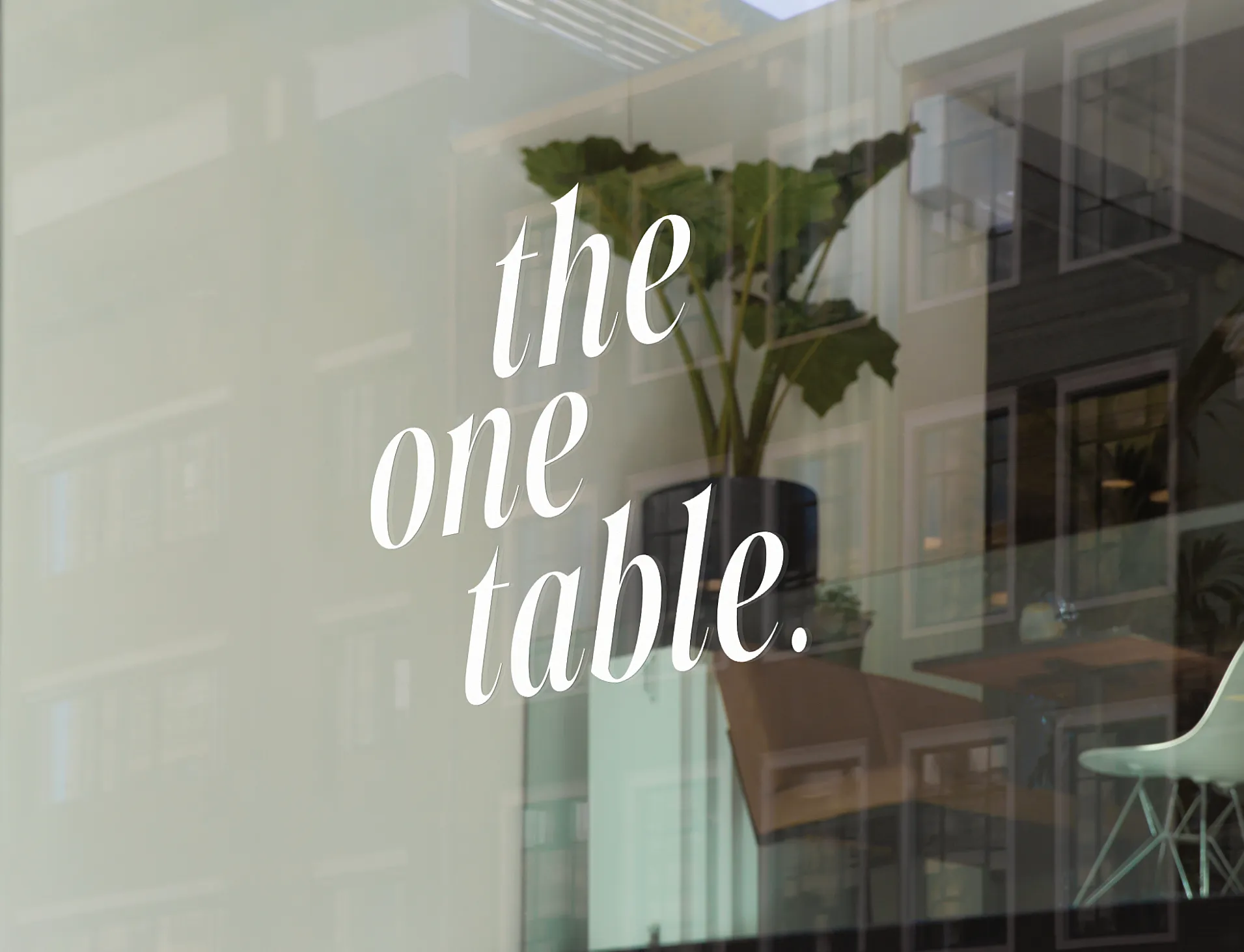Window with white text reading 'the one table.' reflecting indoor plants and chairs inside a modern cafe or restaurant.