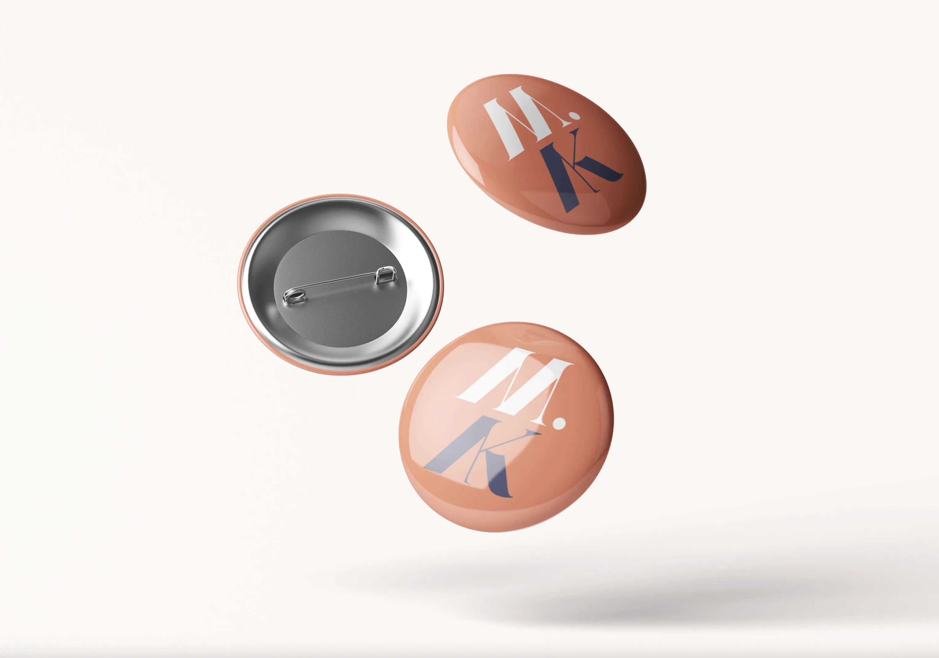 Three round pins with a shiny peach-colored surface and the letters 'M.K' printed on two, one showing the pin back.