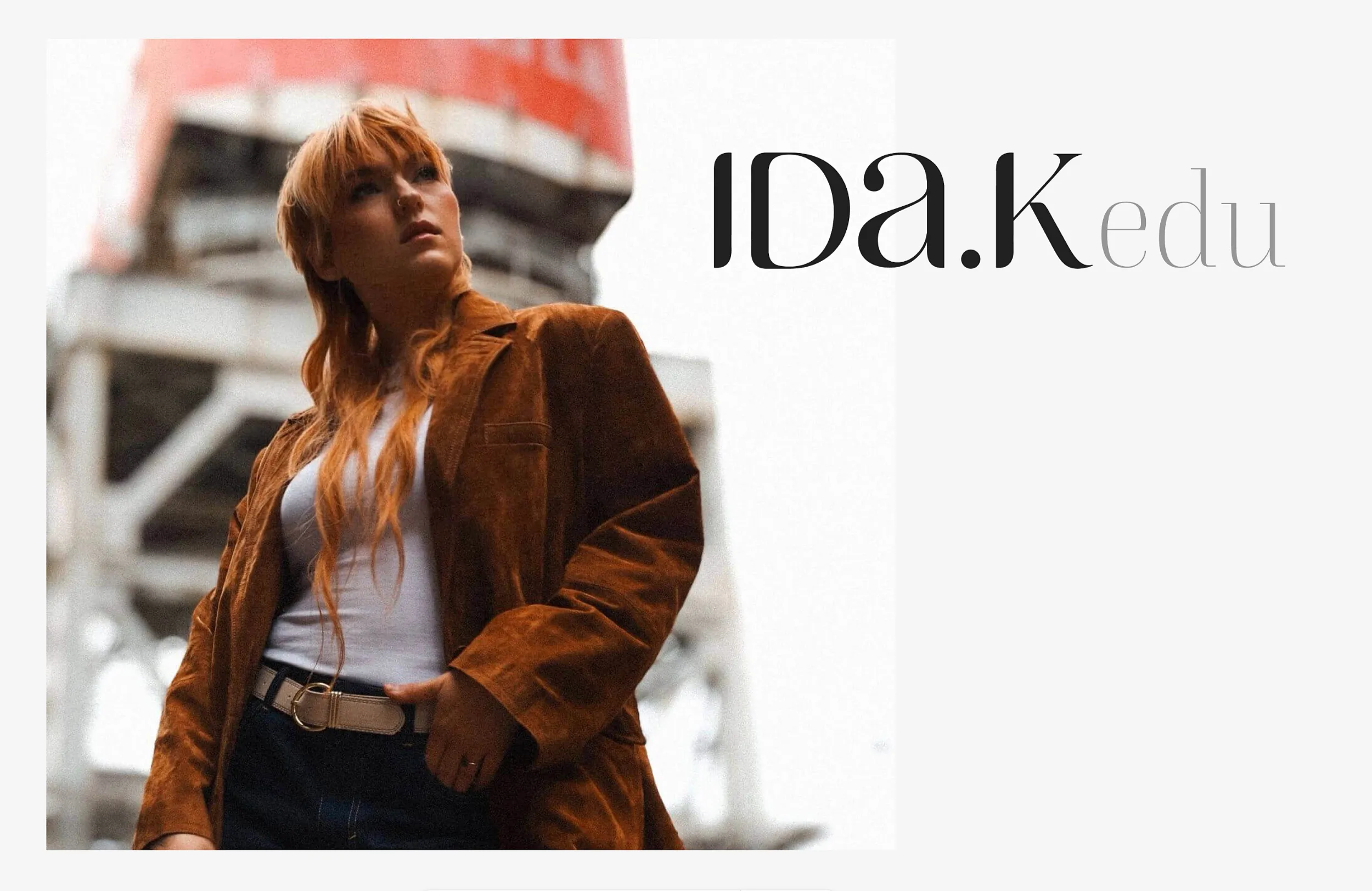 Woman with long red hair wearing a brown jacket and white top standing outdoors with industrial structure behind.