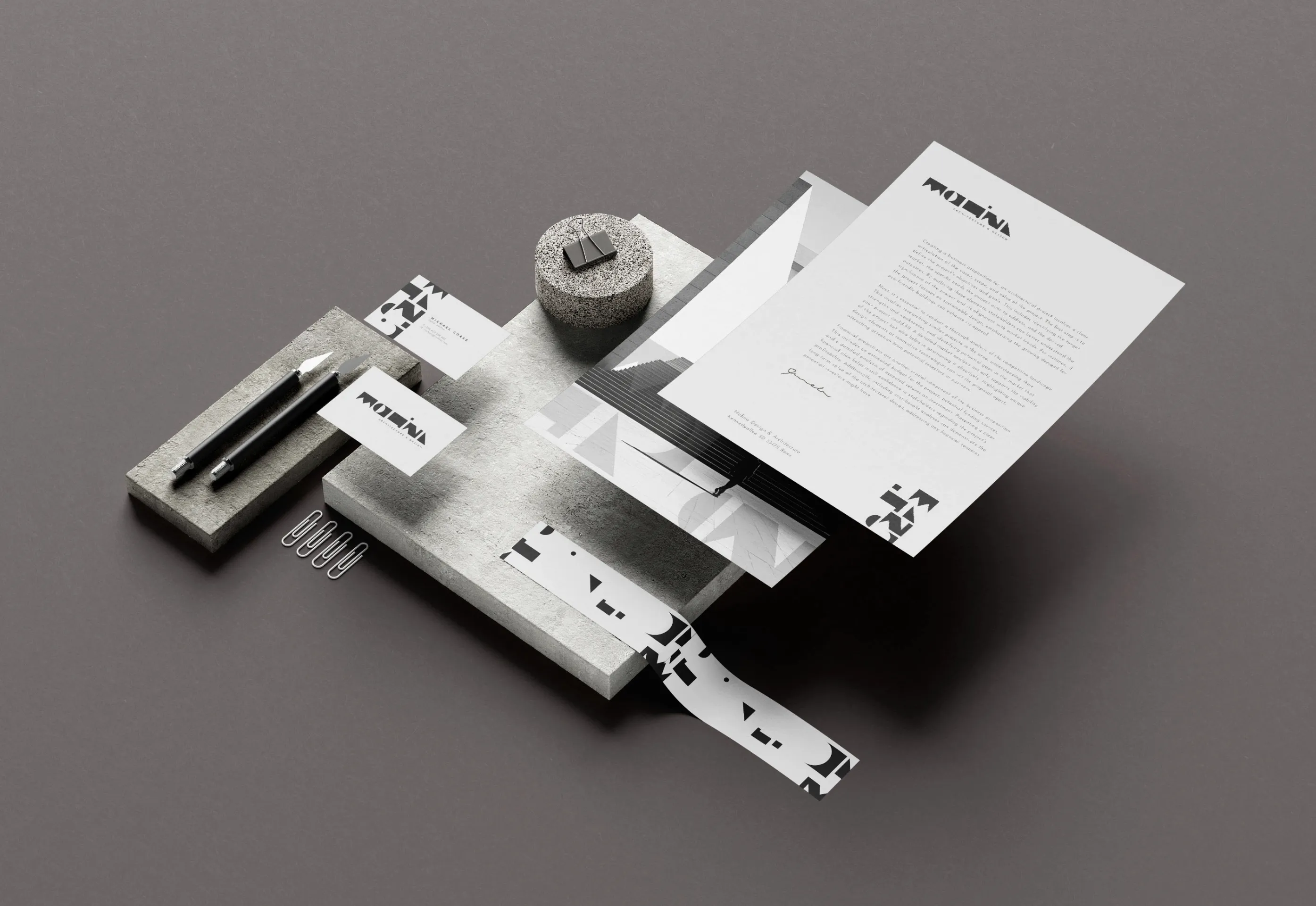 Monochrome branding set including letterhead, business cards, pens, paper clips, and textured blocks arranged on a gray surface.